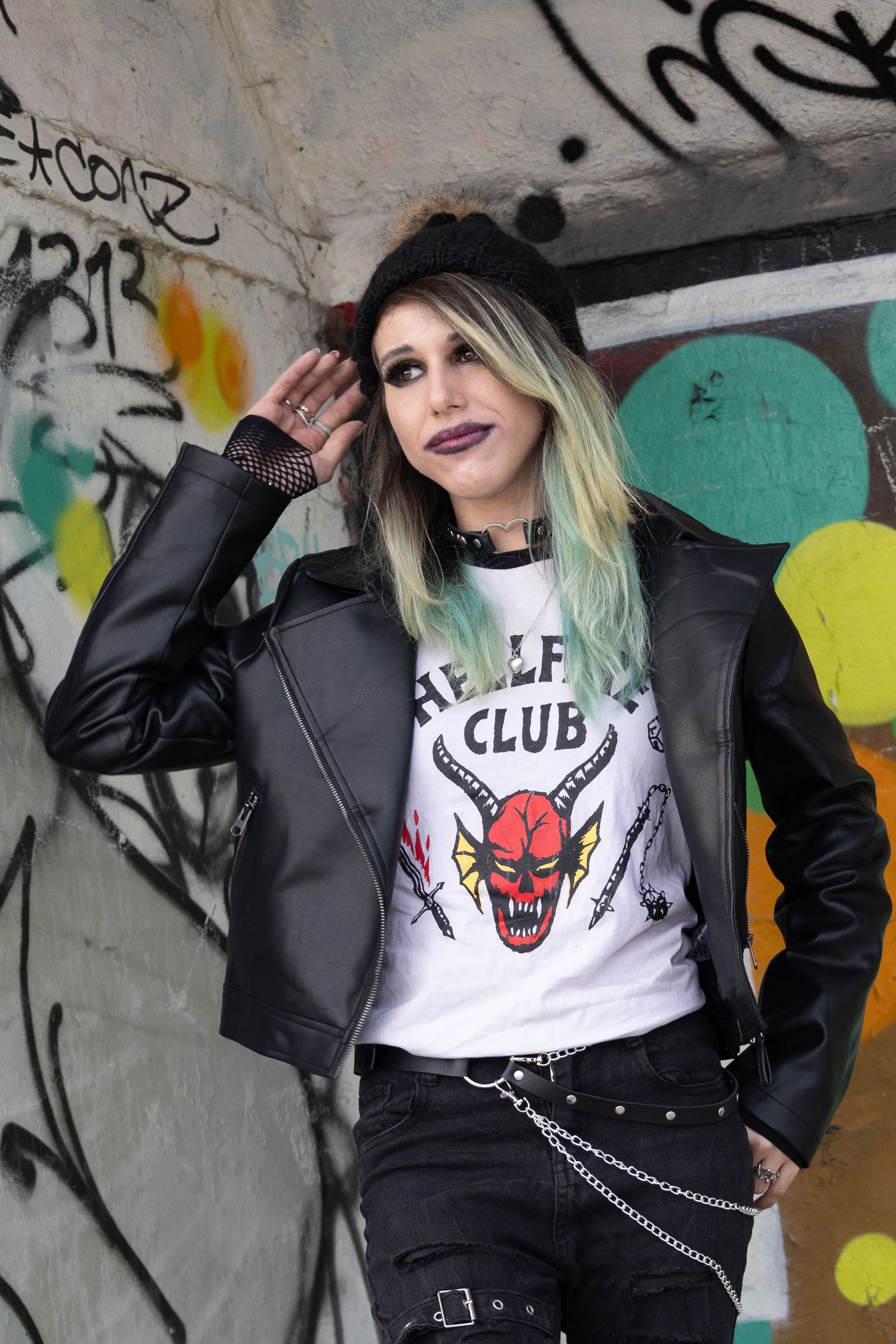 Young woman in punk style wearing Hellfire Club tee, leather jacket, chain belt, posing against graffiti wall.