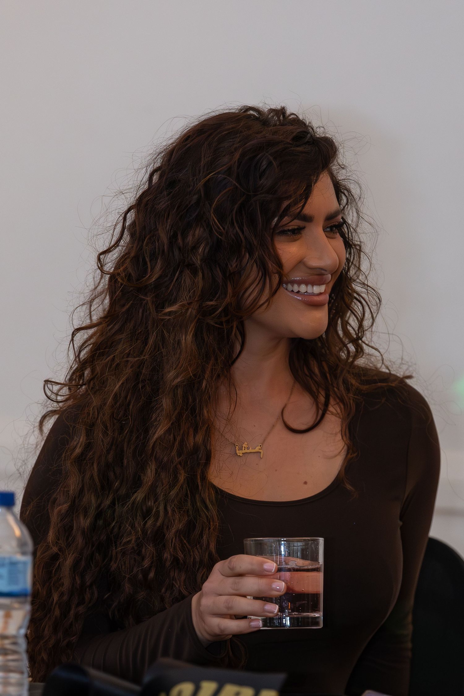 Smiling woman with long curly brown hair holding a glass drink wearing a dark top with gold necklace.