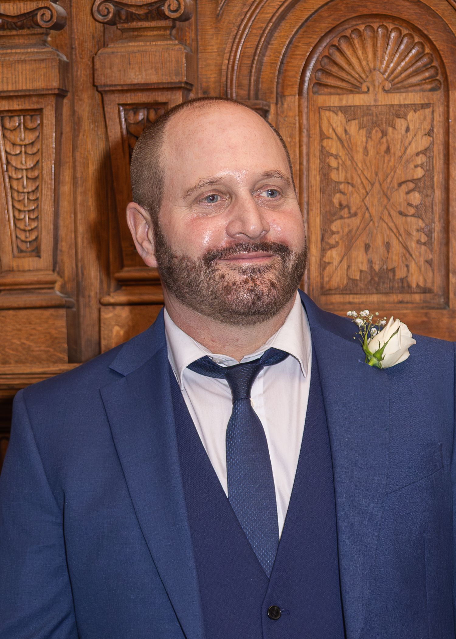 Smiling bearded man in blue suit with boutonniere stands before ornate wooden backdrop at formal event.