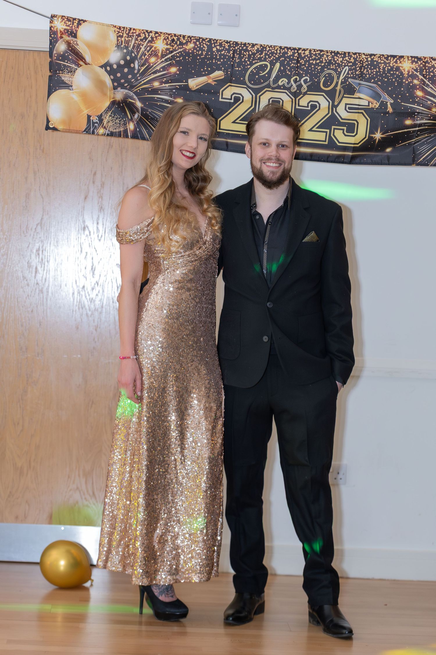 Couple posing at Class of 2025 prom, woman in gold sequin gown and man in black suit with green pocket square.