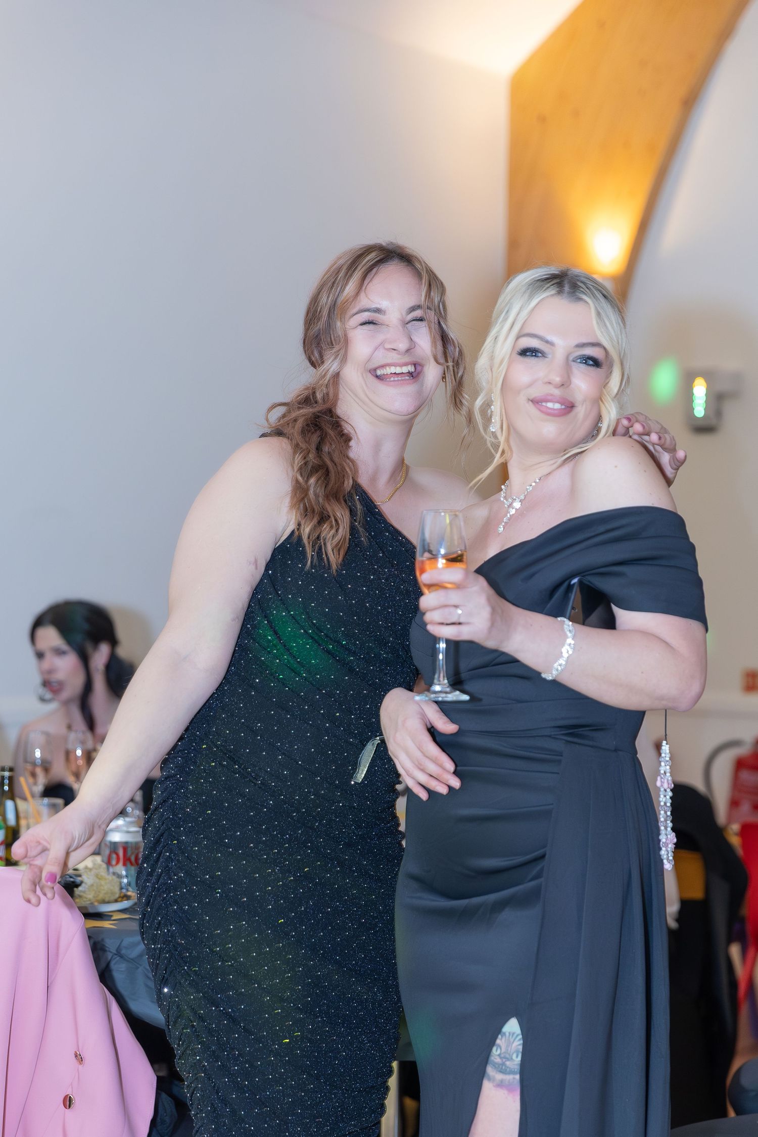 Two women in elegant black gowns smiling and posing together at a formal event, one holding a champagne glass.