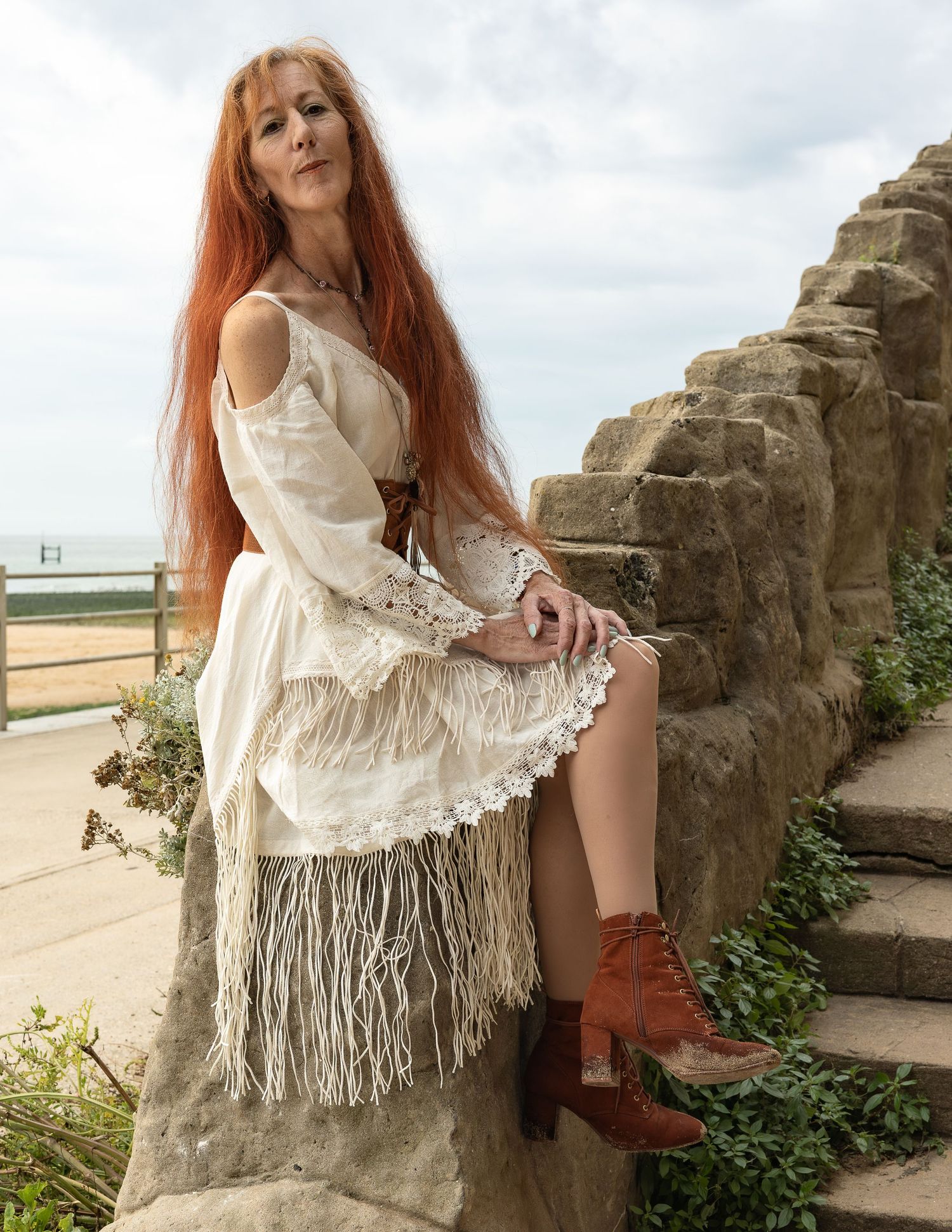 Woman with long red hair in boho white fringe dress and brown boots sitting on stone wall outdoors.