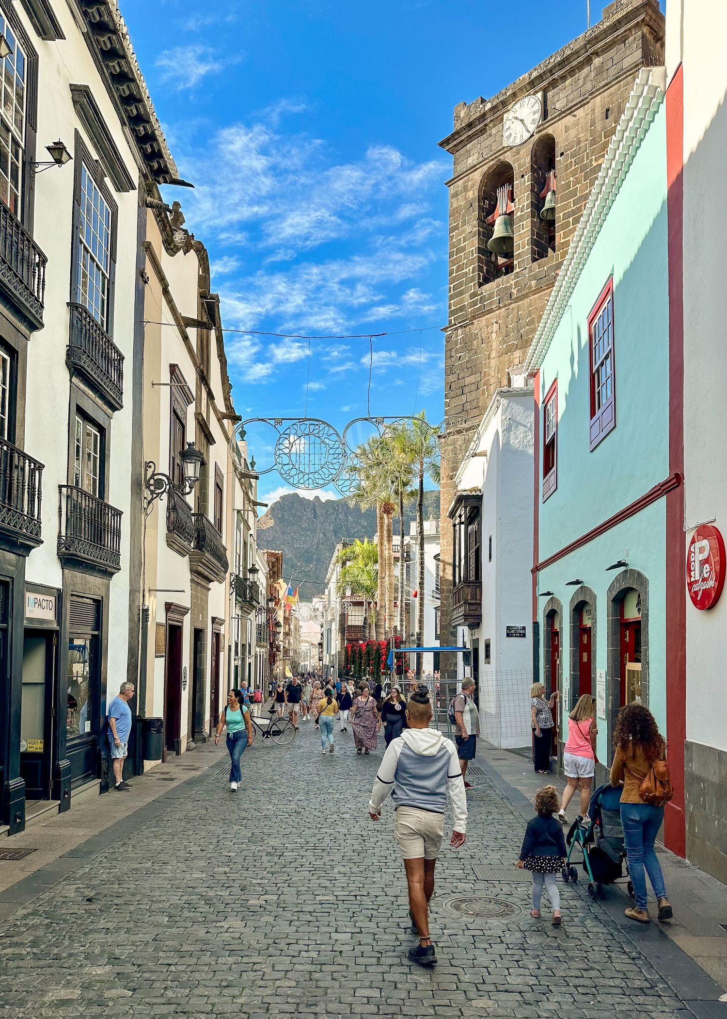 Pedestrians stroll along a charming cobblestone street lined with historic buildings and a church tower in Santa Cruz de La Palma.