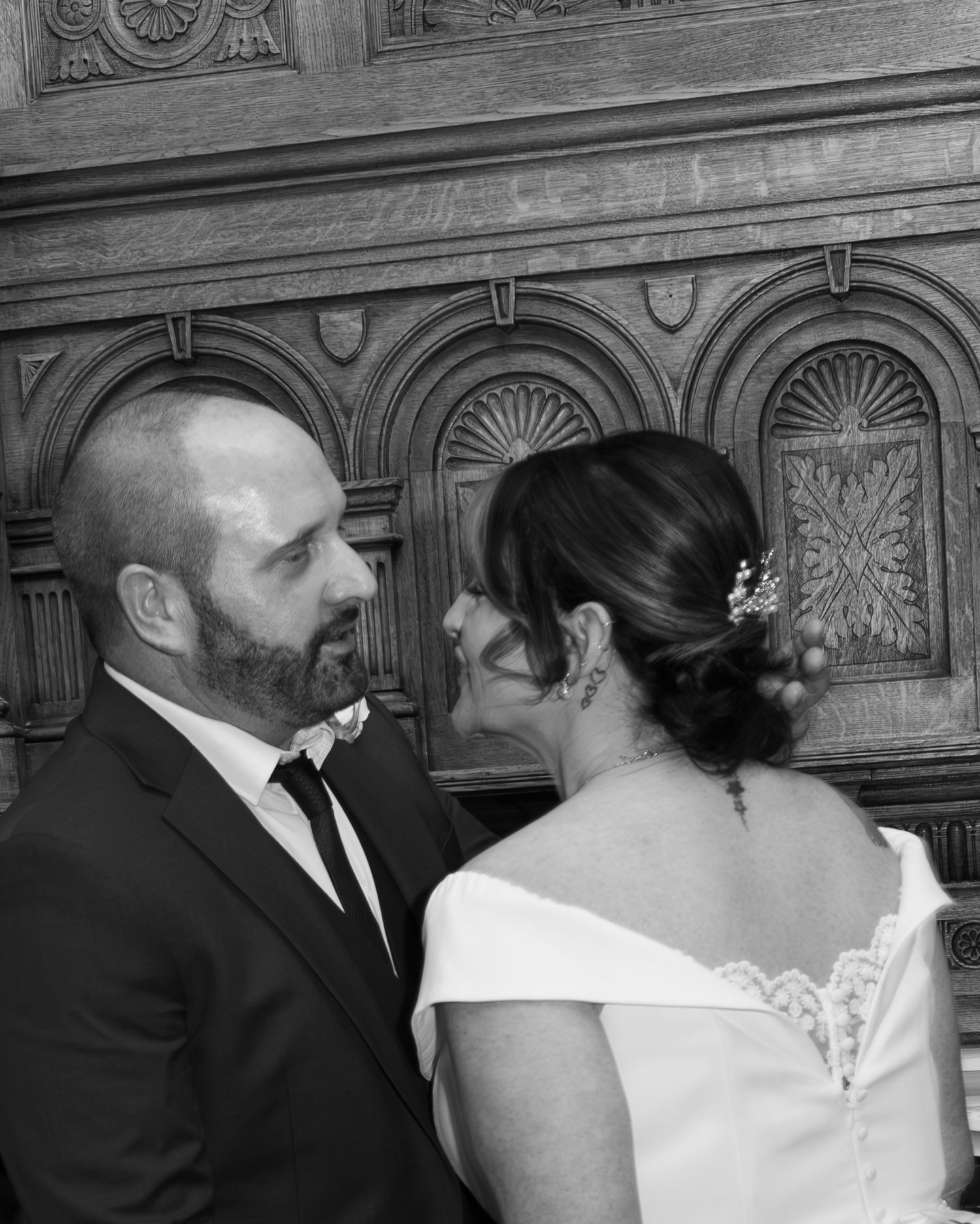 Black and white intimate portrait of bride and groom gazing at each other before ornate wooden backdrop.