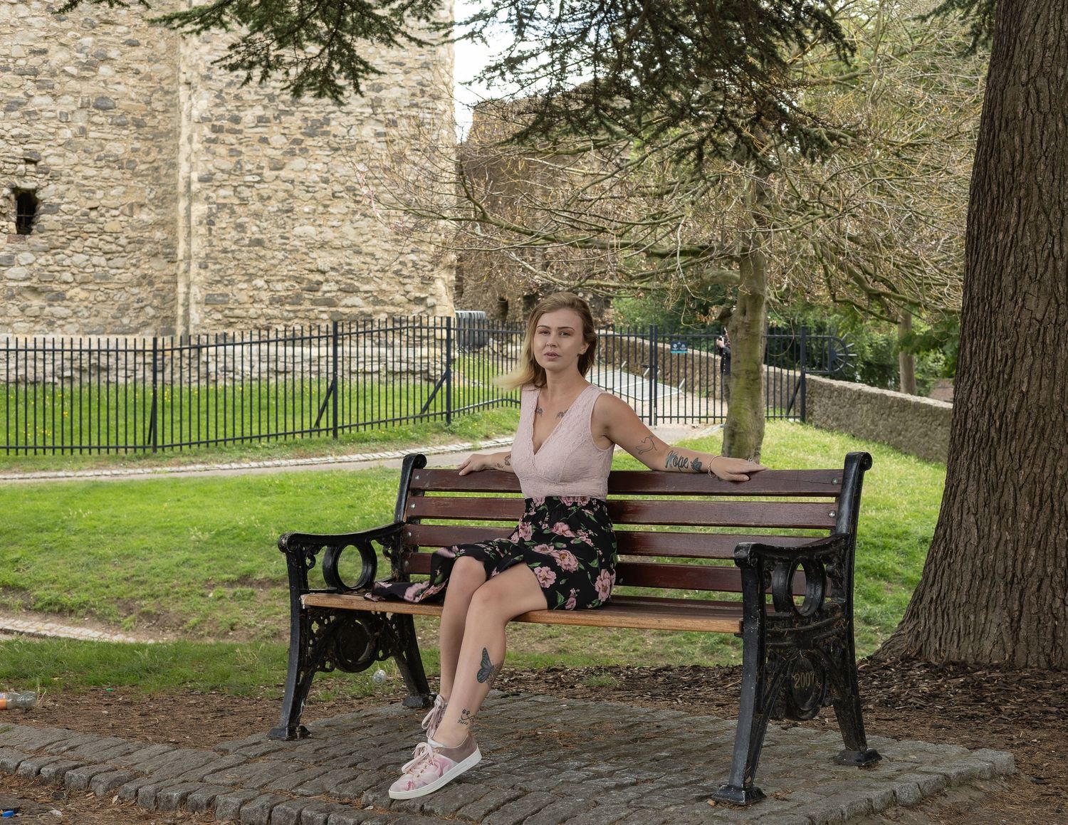 Woman in pink top and floral skirt sitting on a wooden park bench near historic stone castle walls.
