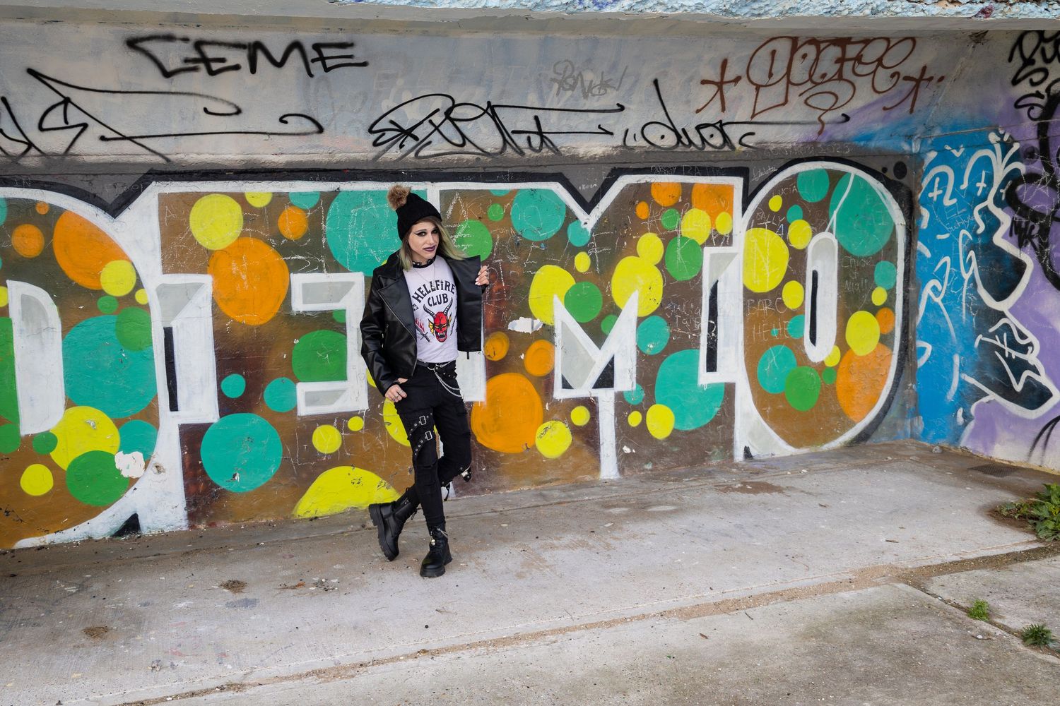 Person posing against a colorful graffiti mural wall with green, orange, and teal circle designs outdoors.