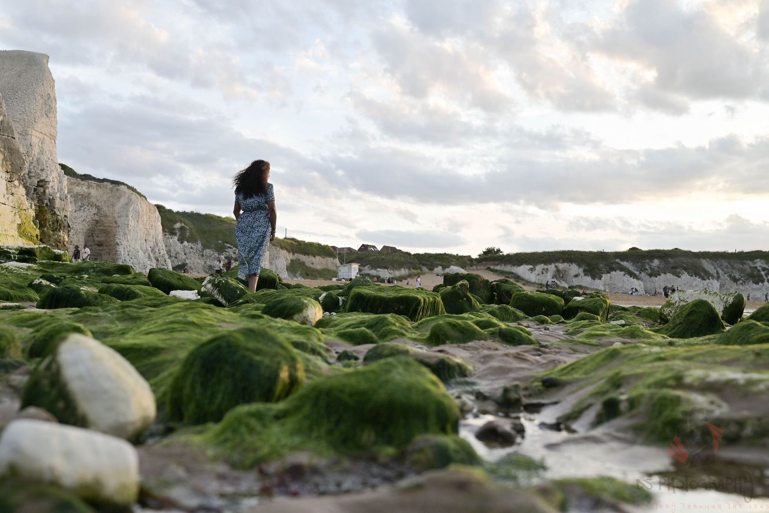 Woman in a dress walks along a rocky, moss-covered beach with white cliffs under a cloudy sky.