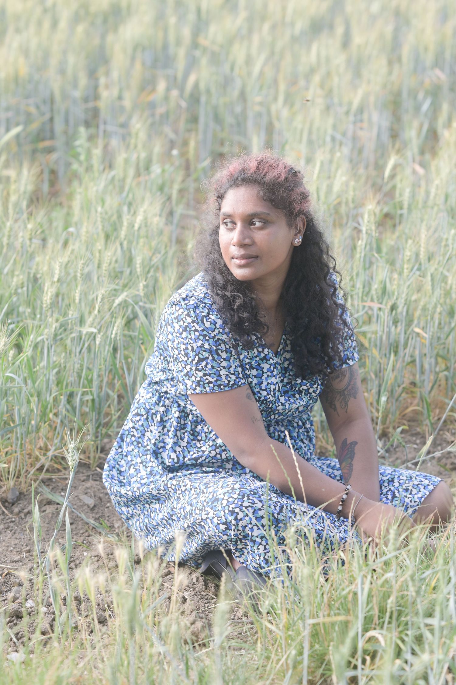 A young woman with curly hair sits gracefully in a green field, wearing a blue floral dress, gazing into the distance.
