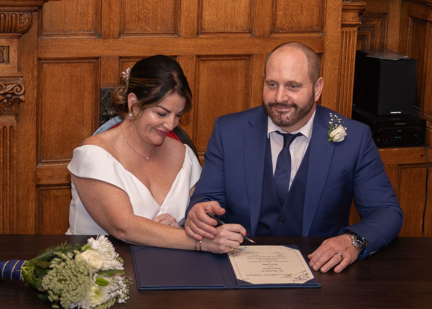 Couple smiling and signing wedding register at ceremony table with bridal bouquet in elegant wood-paneled room.