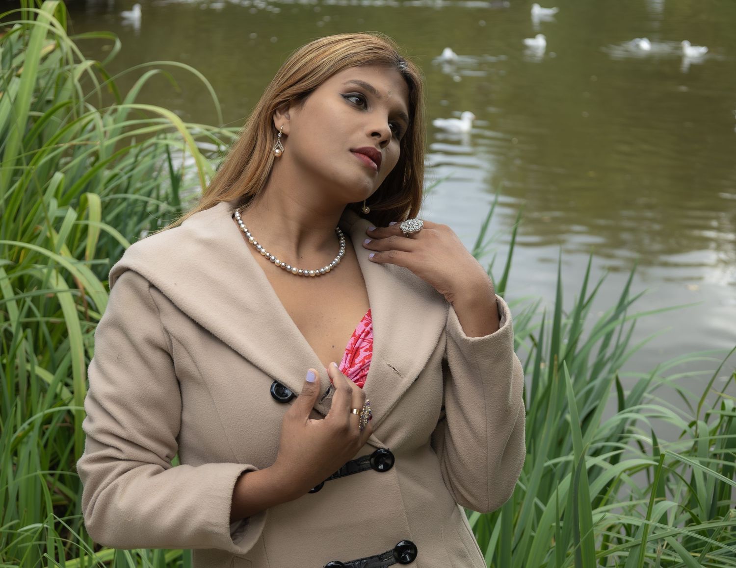 Woman in beige blazer and pearl necklace poses near a pond with geese and lush green reeds.