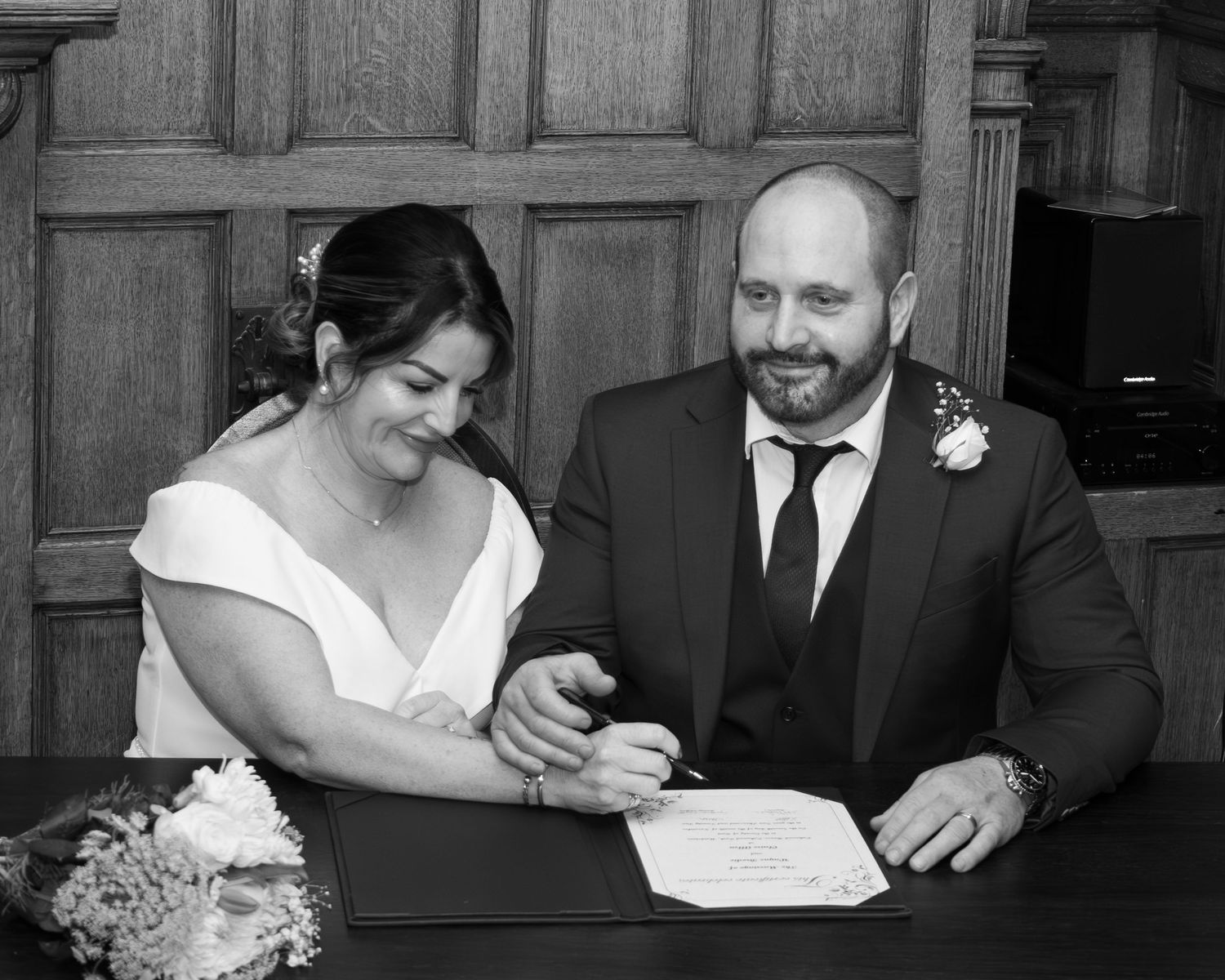 Black and white photo of a smiling couple signing their marriage certificate at a wooden desk, bride holding flowers.
