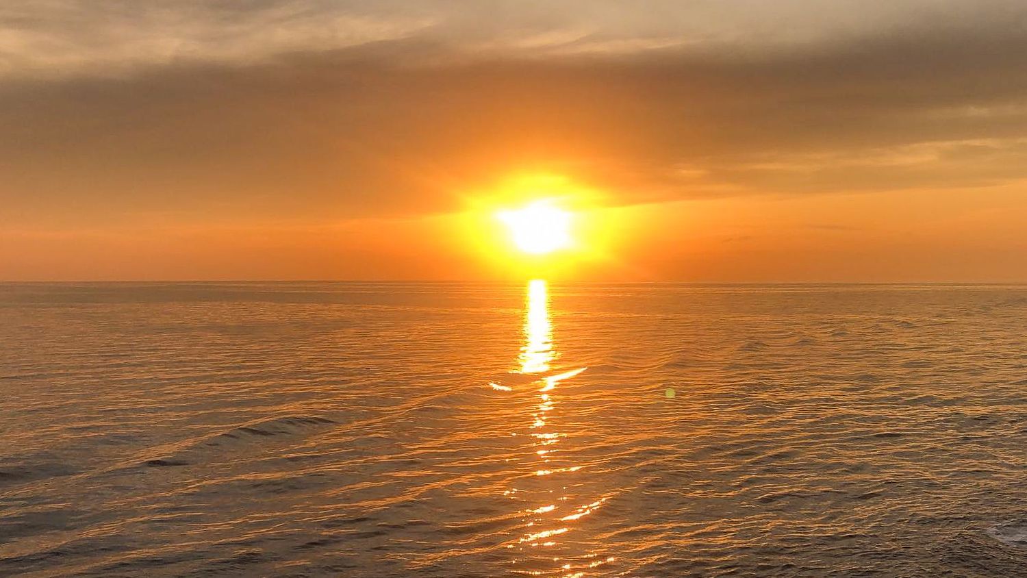Golden sunset over calm ocean waters with sun reflecting on gentle waves under a dramatic orange sky.