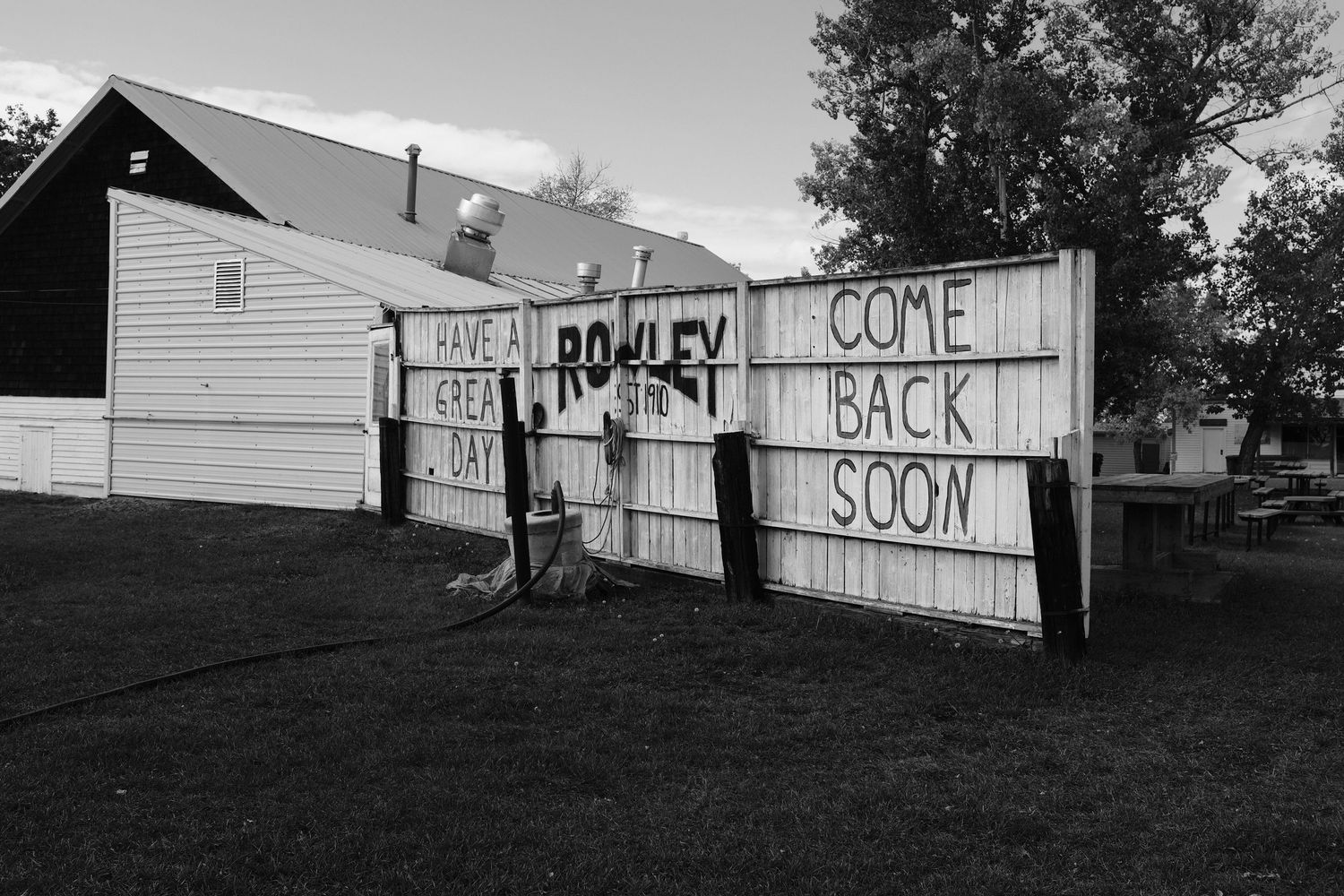 Black and white photo of a fence sign reading Rowley Come Back Soon with Have a Great Day in a small town.