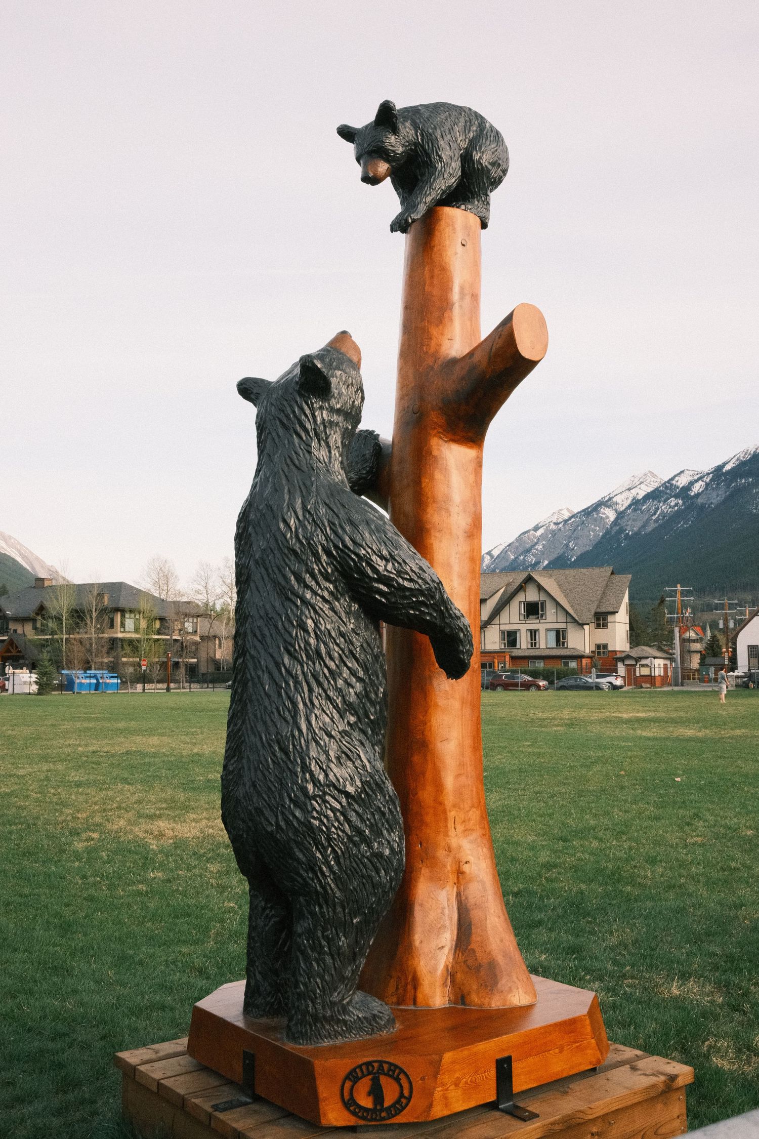 Bronze bear sculpture with cub climbing wooden tree trunk, set against mountain backdrop in small town park.