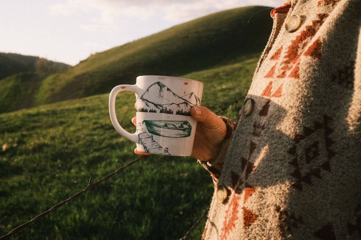 Person holding a white camping mug with mountain design in a green meadow, wearing a cozy Aztec-print blanket.