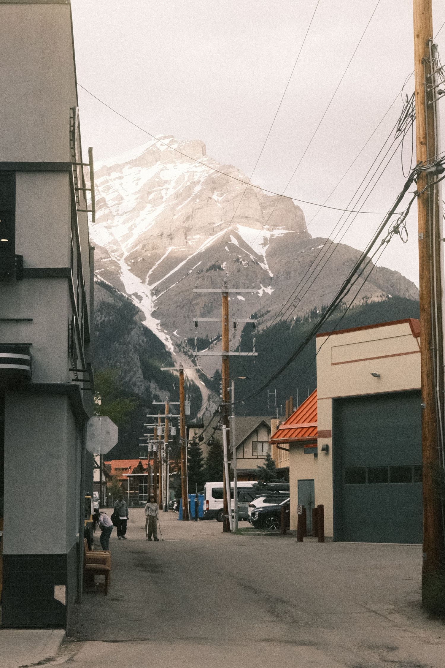 A small mountain town street with snow-capped peaks, utility poles, and buildings under an overcast sky.
