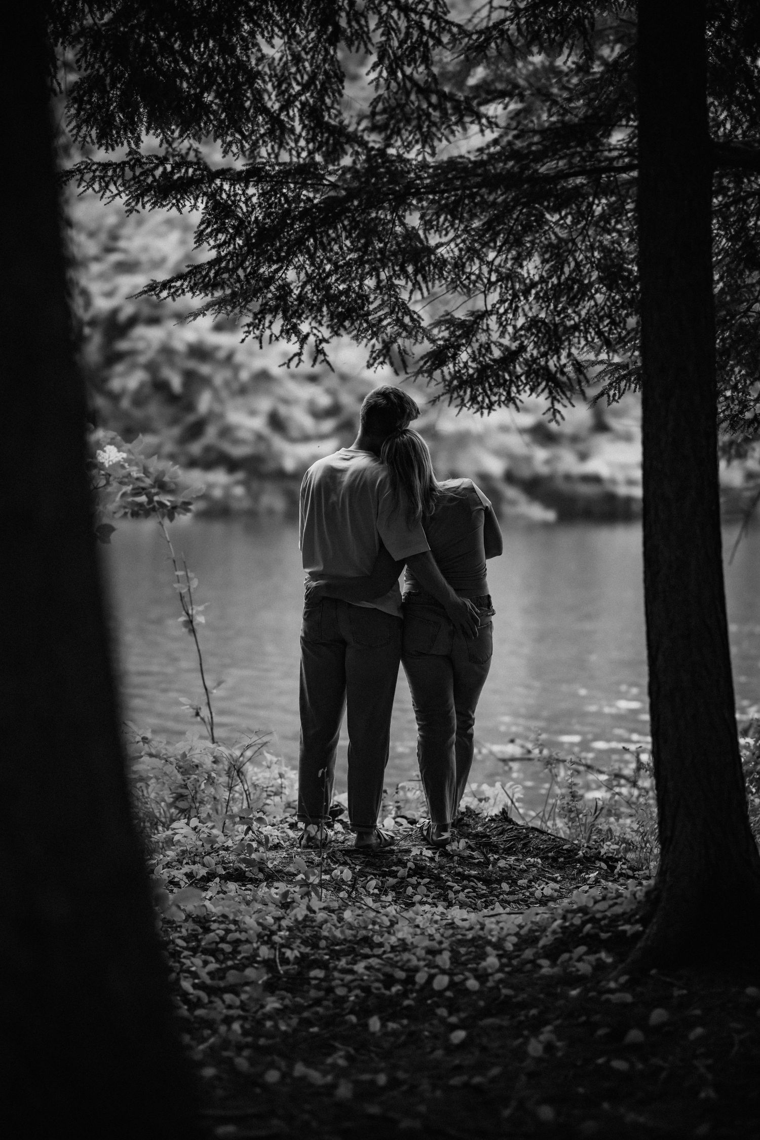 Silhouetted couple standing together by lake through trees in black and white.