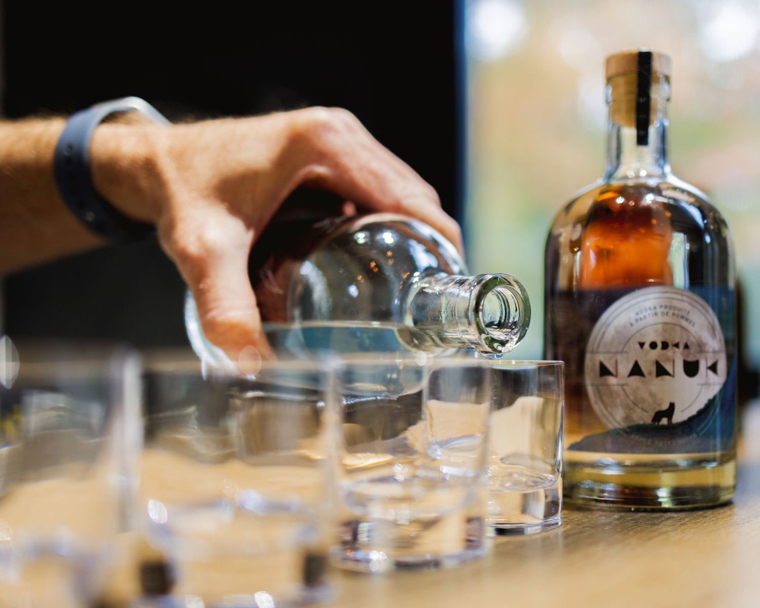 Row of clear glasses on a bar counter with bottles of spirits in the background.