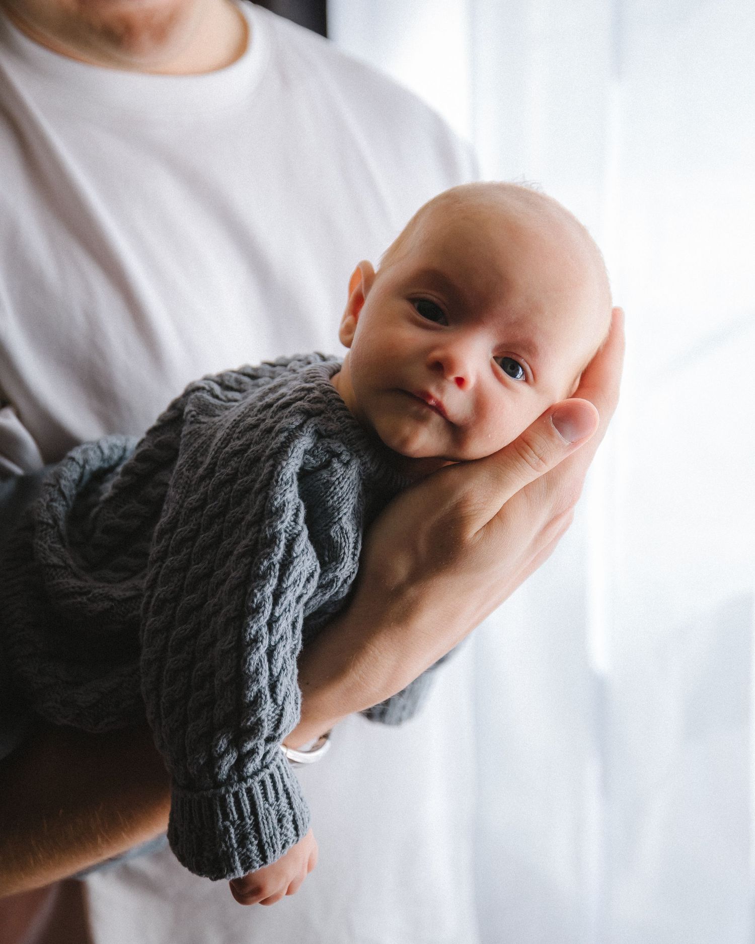 Newborn baby in gray knit sweater being held against white background.