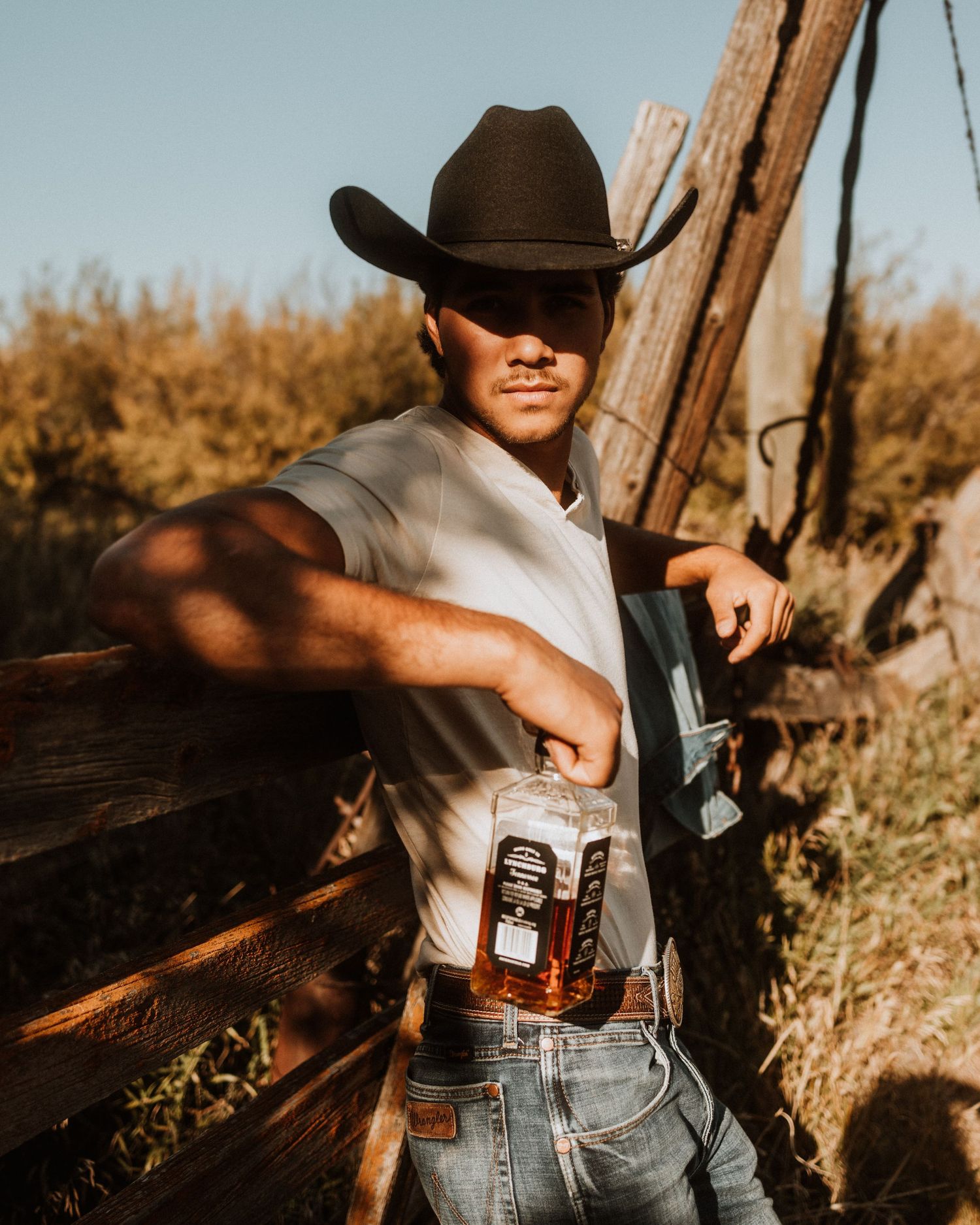 Cowboy in white shirt and hat leans against wooden post in golden sunlight.