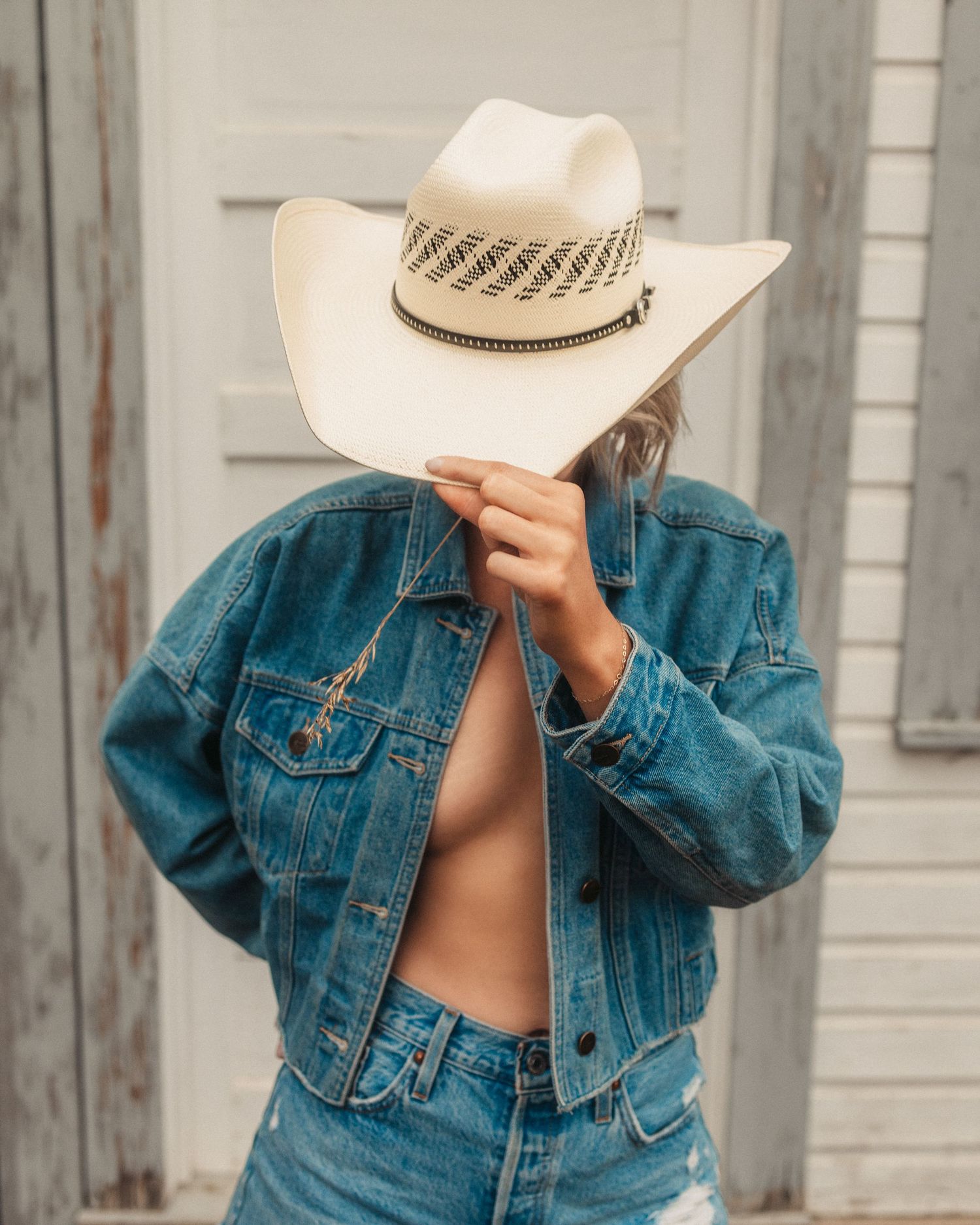 Woman in denim jacket and white cowboy hat poses against rustic wooden wall.