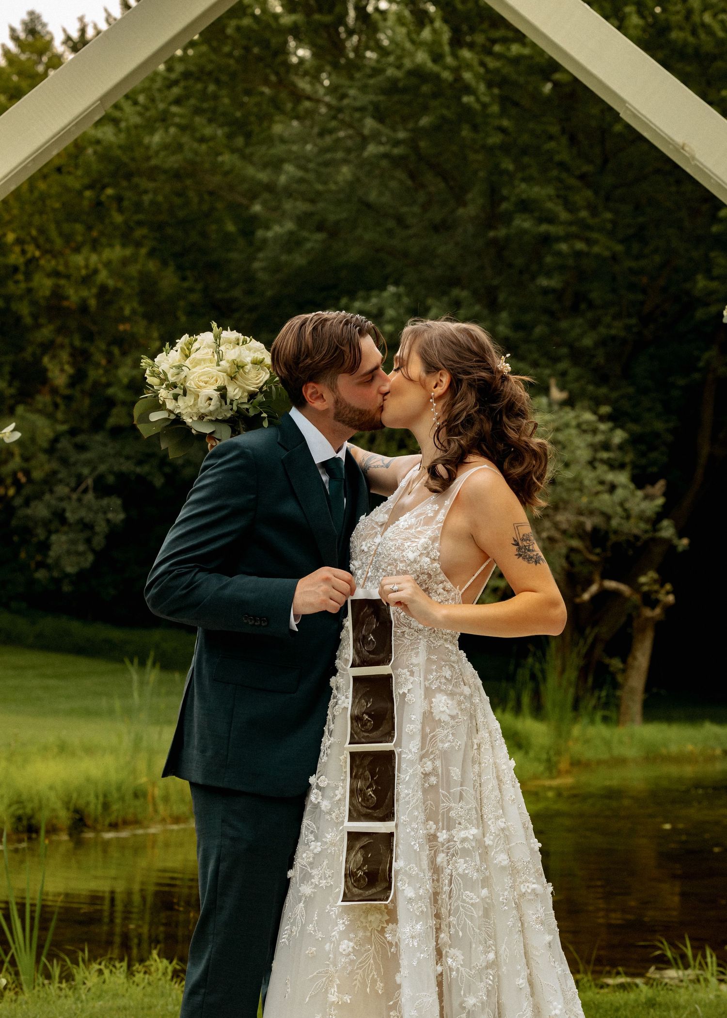 Newlyweds share an intimate moment under a white pergola by a scenic pond.