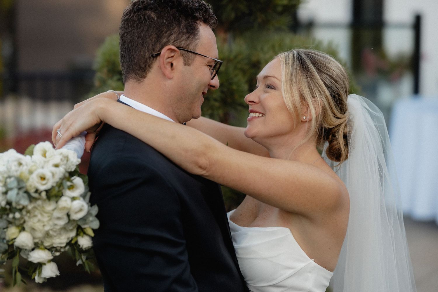 Candid moment of newlyweds embracing outdoors with white floral arrangements.