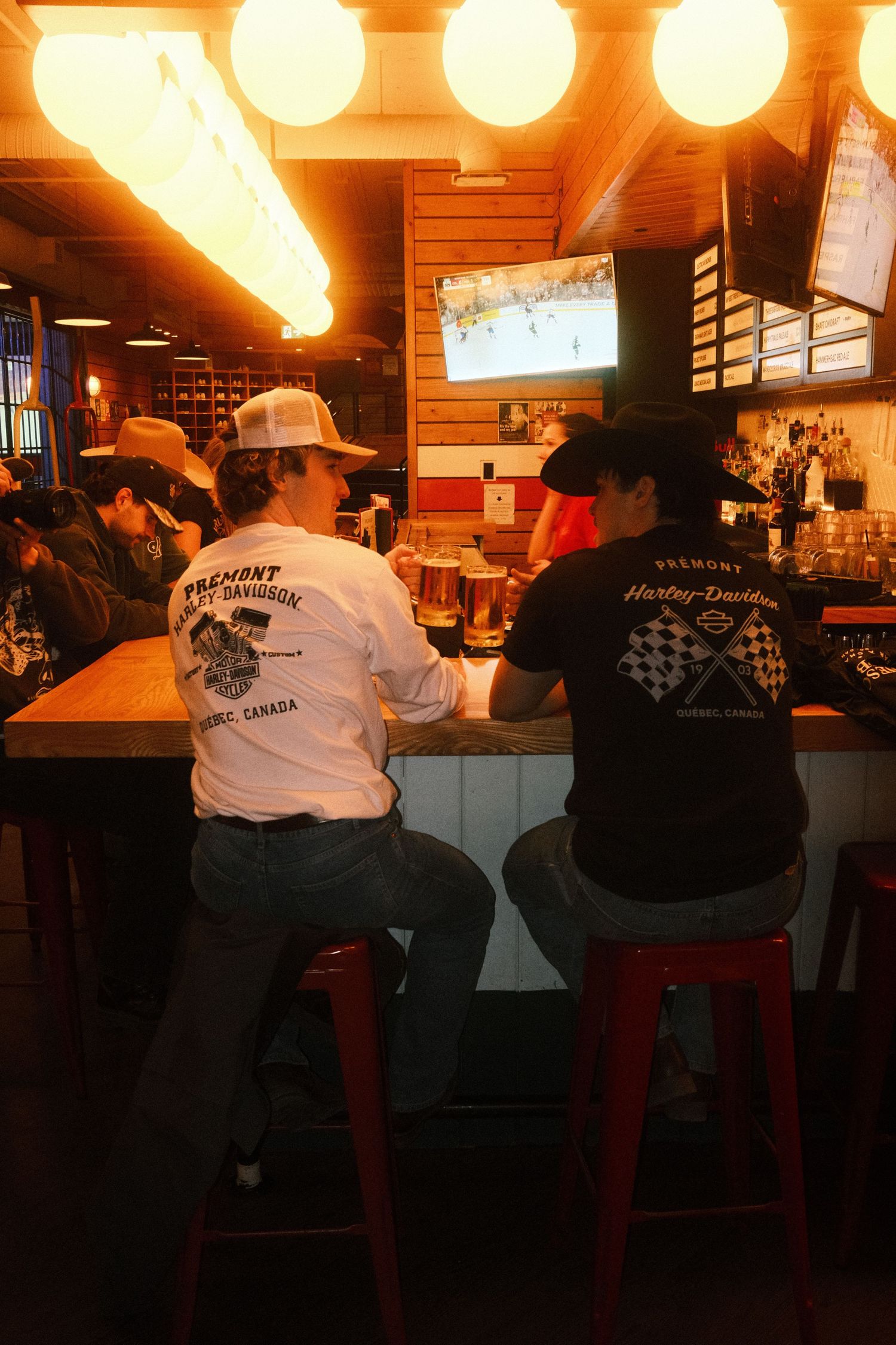 Two people wearing Fremont Harley-Davidson shirts and cowboy hats sit at a rustic bar watching TV.
