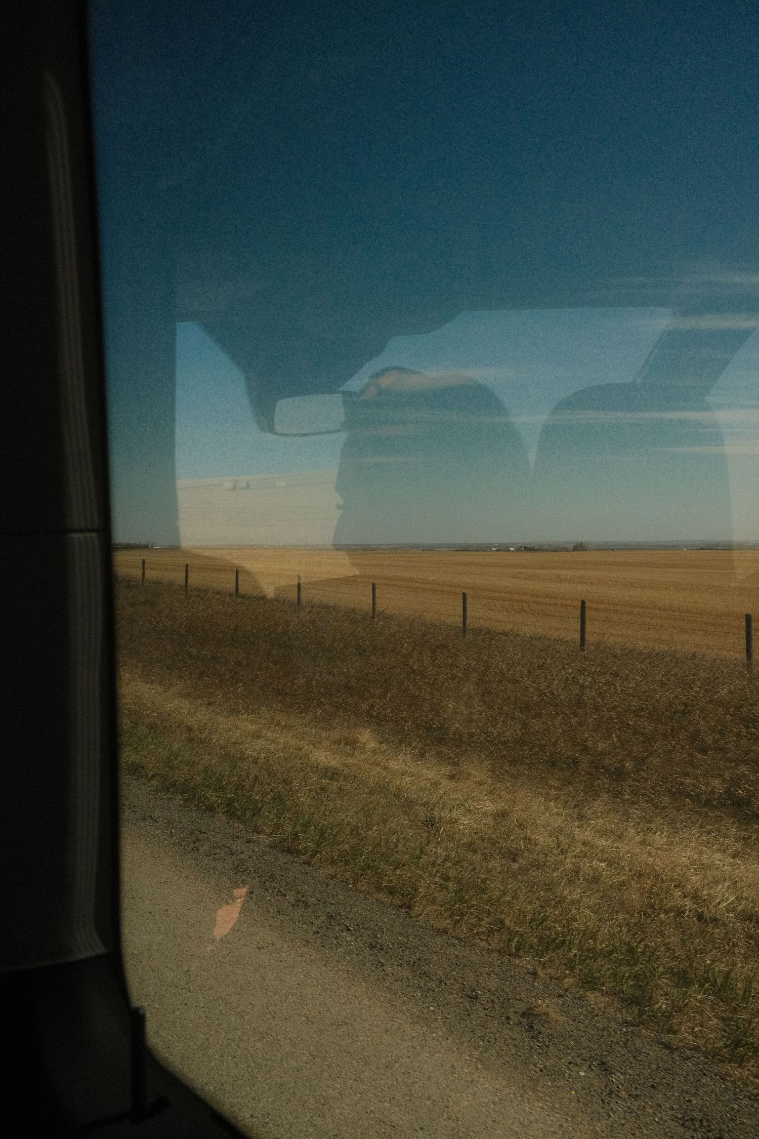 View from a vehicle window of flat rural farmland with fence posts under a clear blue sky.
