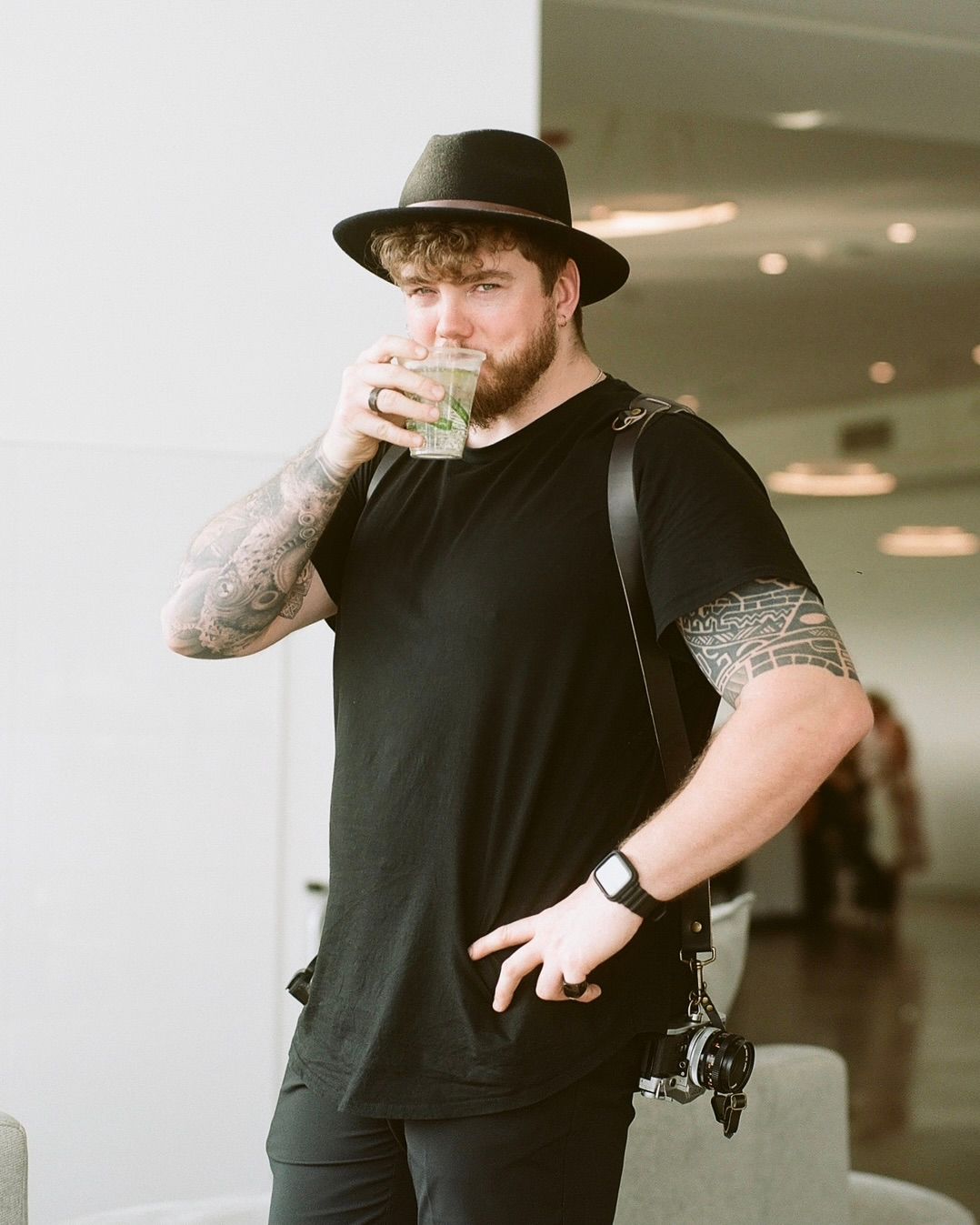 Tattooed man in black outfit and wide-brim hat drinking iced beverage while carrying camera bag indoors.