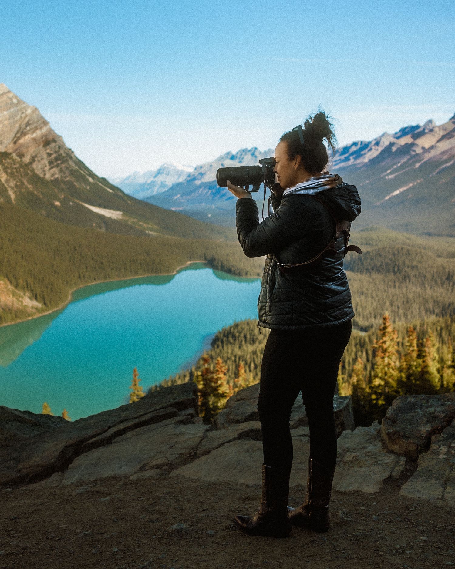 Woman photographing turquoise alpine lake surrounded by mountains and pine forests from rocky overlook.