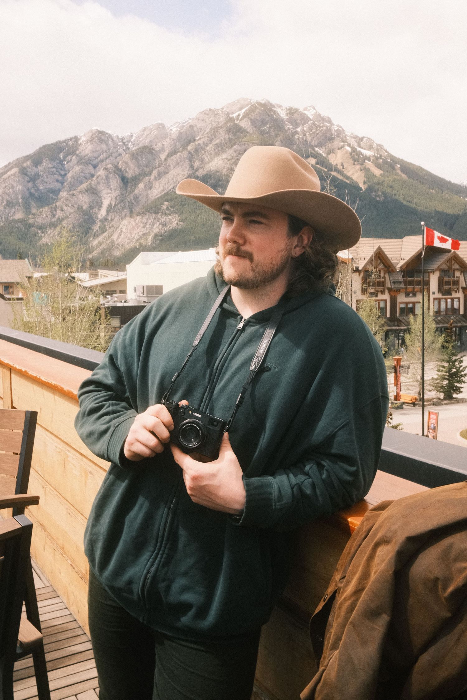 Man in cowboy hat holding camera on balcony with Canadian Rockies mountain backdrop and Canadian flag.