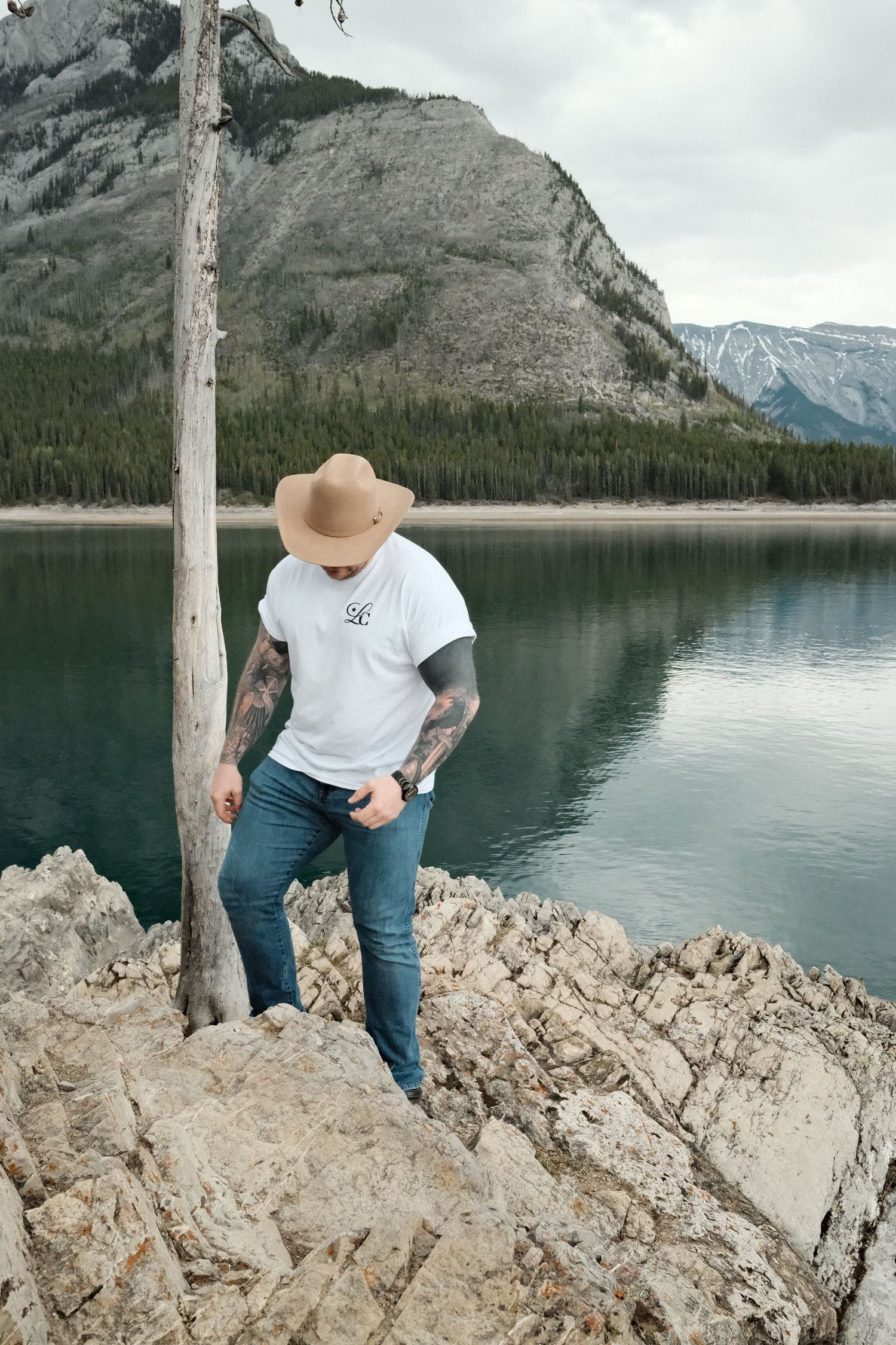 Tattooed man in cowboy hat and white tee stands on rocky shore by turquoise mountain lake with forested peaks behind.