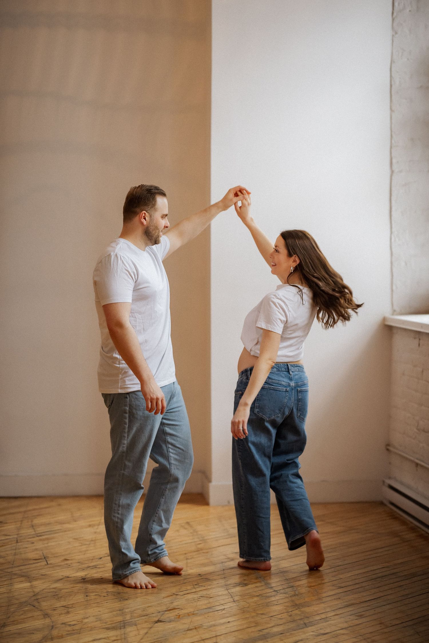 Two people in white shirts and jeans dancing playfully on hardwood floor.