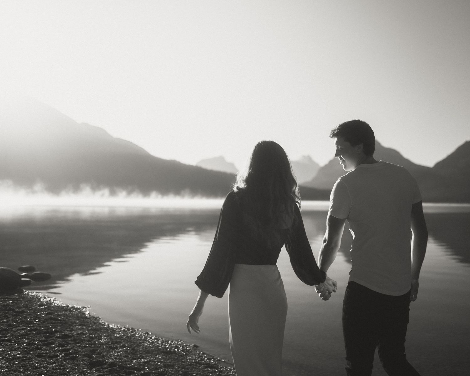 Silhouetted couple standing by a serene mountain lake at sunset.