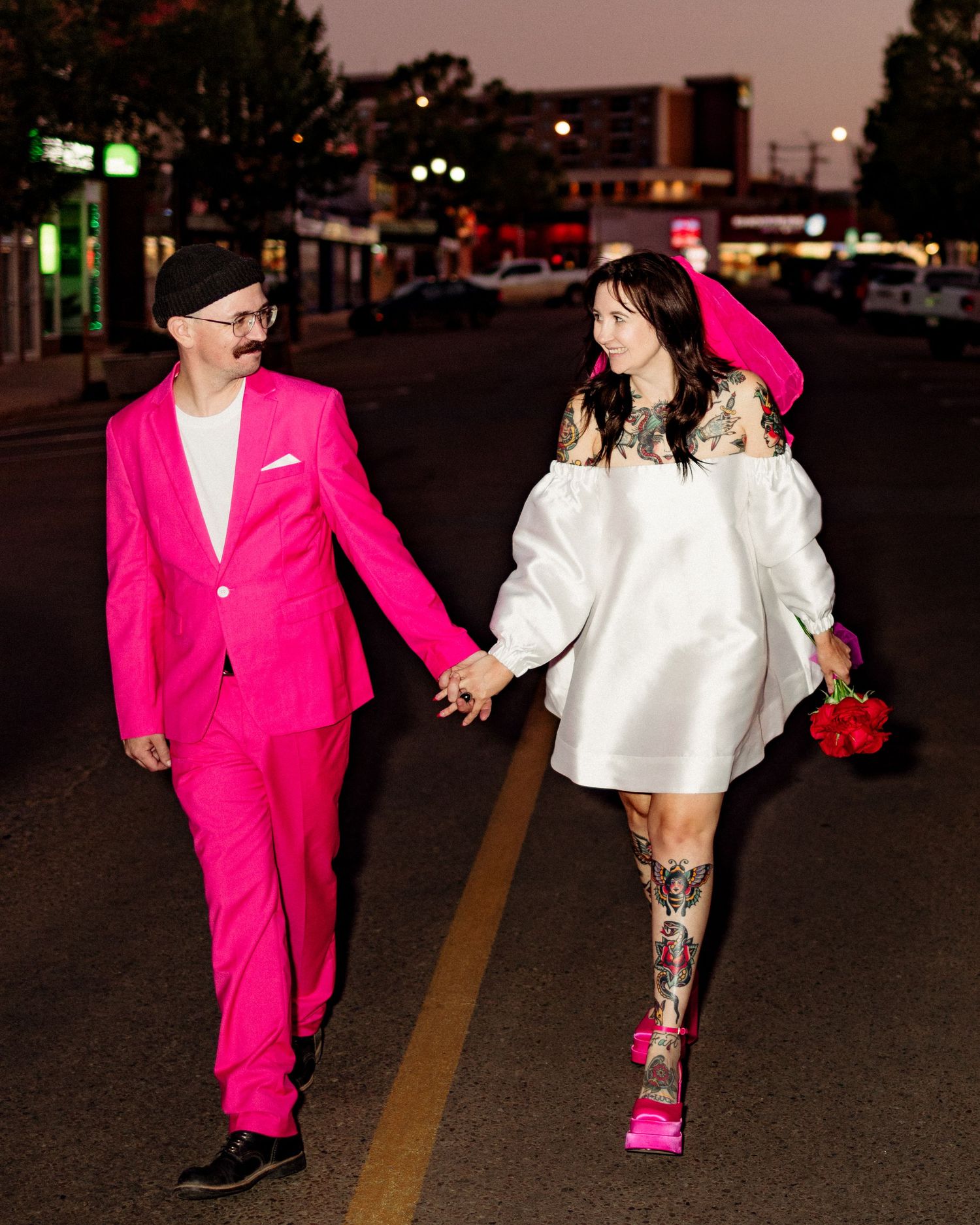 Couple in pink suit and white dress walking playfully on a nighttime street.