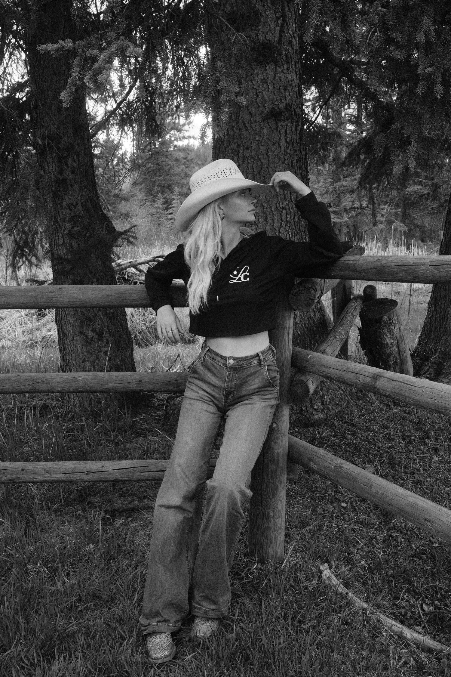 Black and white photo of a blonde woman in cowboy hat, crop top, and flare jeans leaning on a rustic wooden fence.