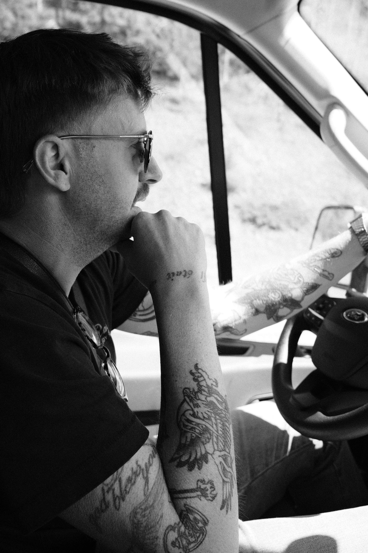 Black and white photo of tattooed man wearing sunglasses sitting in driver seat of vehicle.