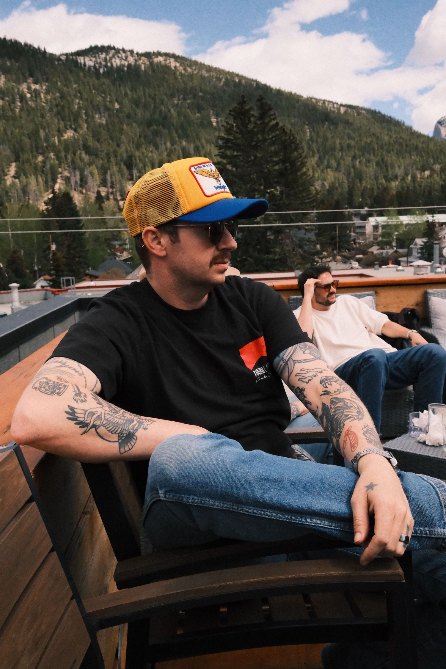 Tattooed man in yellow trucker hat and sunglasses relaxing on rooftop deck with mountain backdrop.