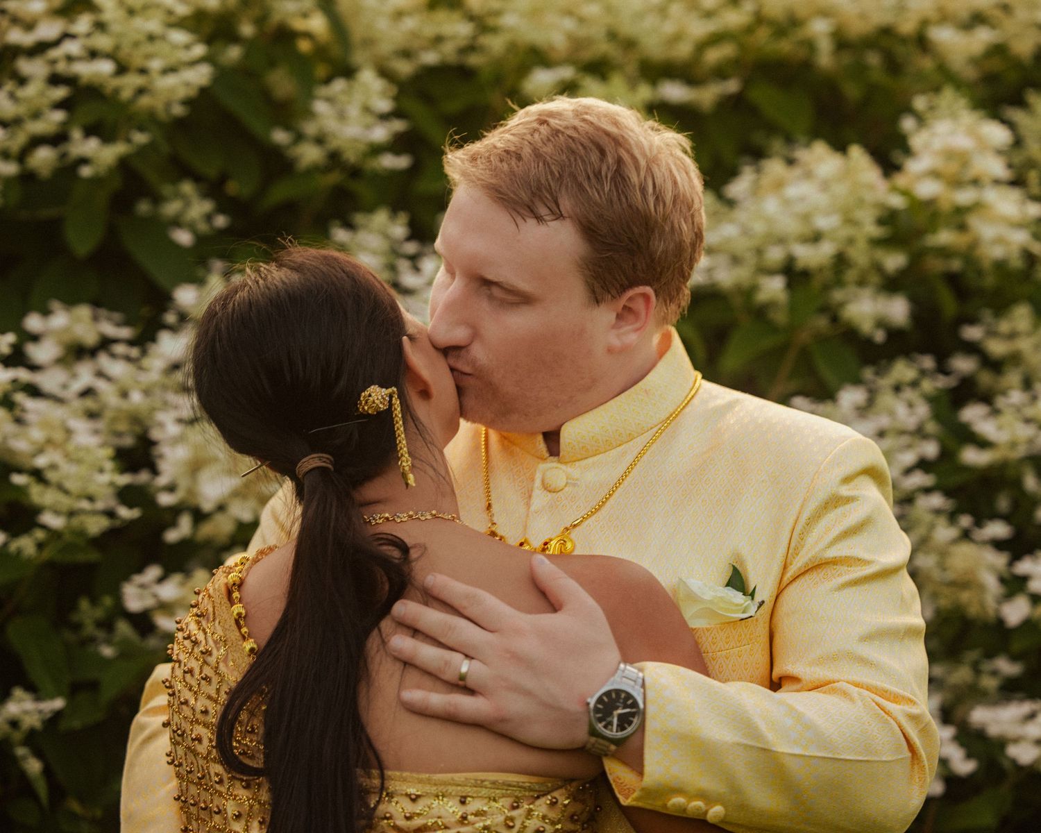 A couple in yellow traditional attire share an intimate embrace against a backdrop of white flowers.