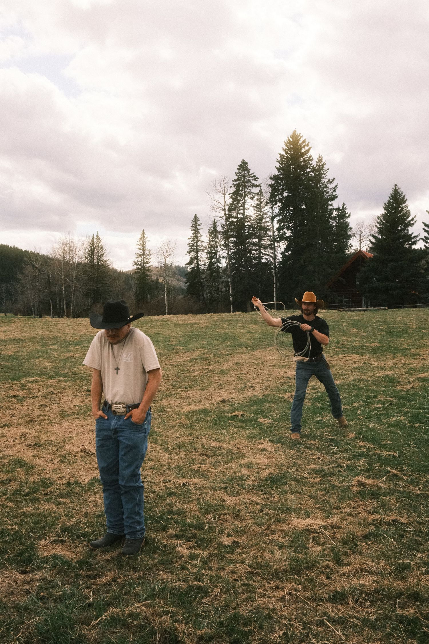 Two men in cowboy hats and jeans stand in a rural field with pine trees, one swinging a baseball bat.