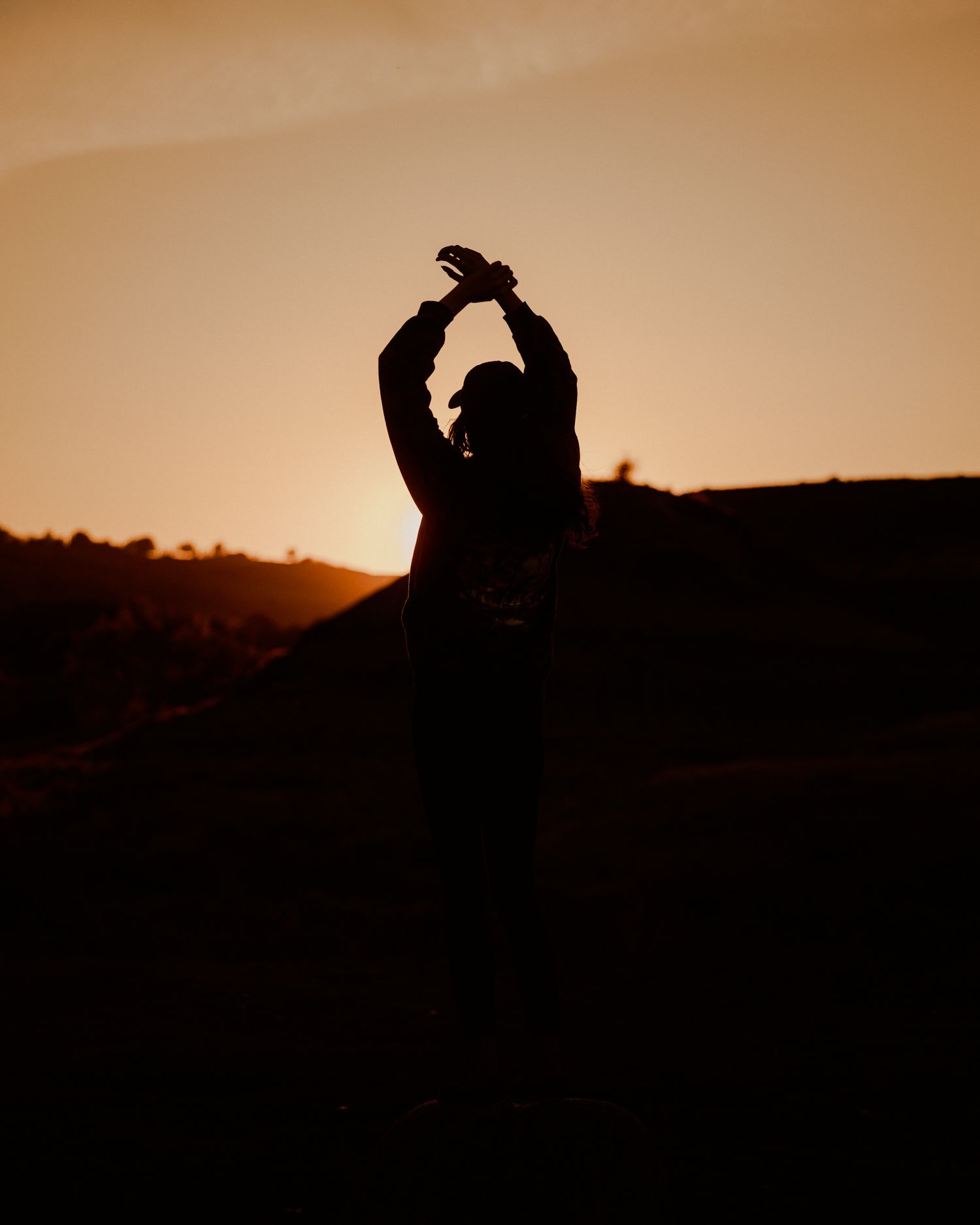 Silhouette of a person dancing with arms raised against a warm golden sunset sky outdoors.