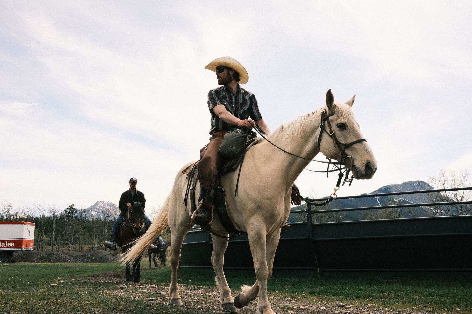 A cowgirl in a hat rides a white horse at a ranch with mountains in the background.