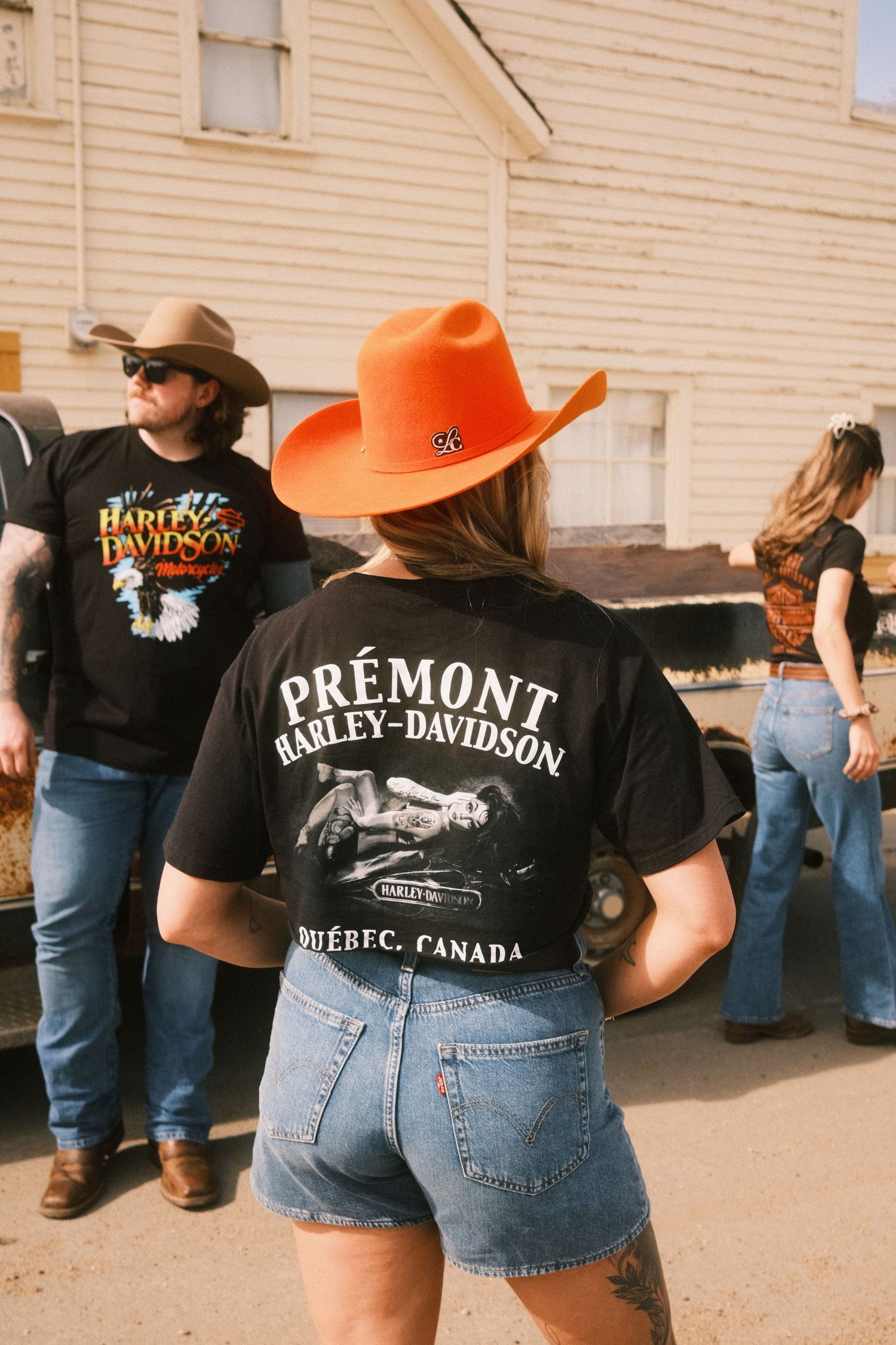 Woman in orange cowboy hat and Prémont Harley-Davidson Québec black tee with denim shorts at outdoor gathering.