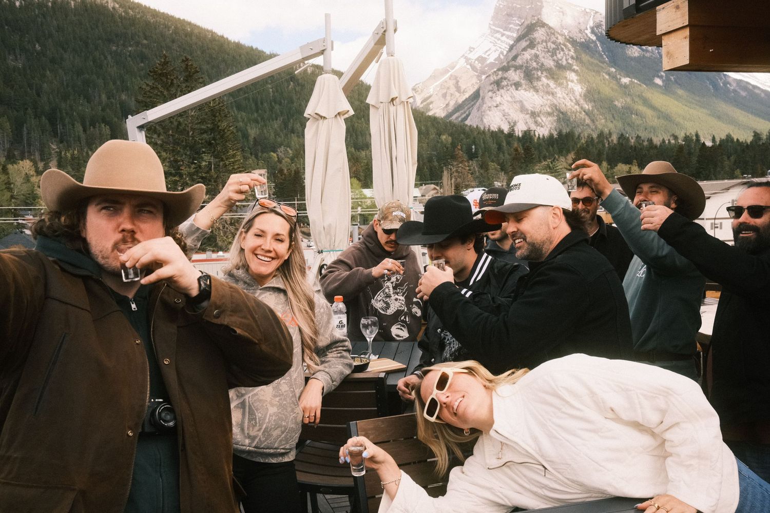 Group of friends celebrating outdoors on a mountain deck, raising glasses and laughing with scenic peaks behind them.
