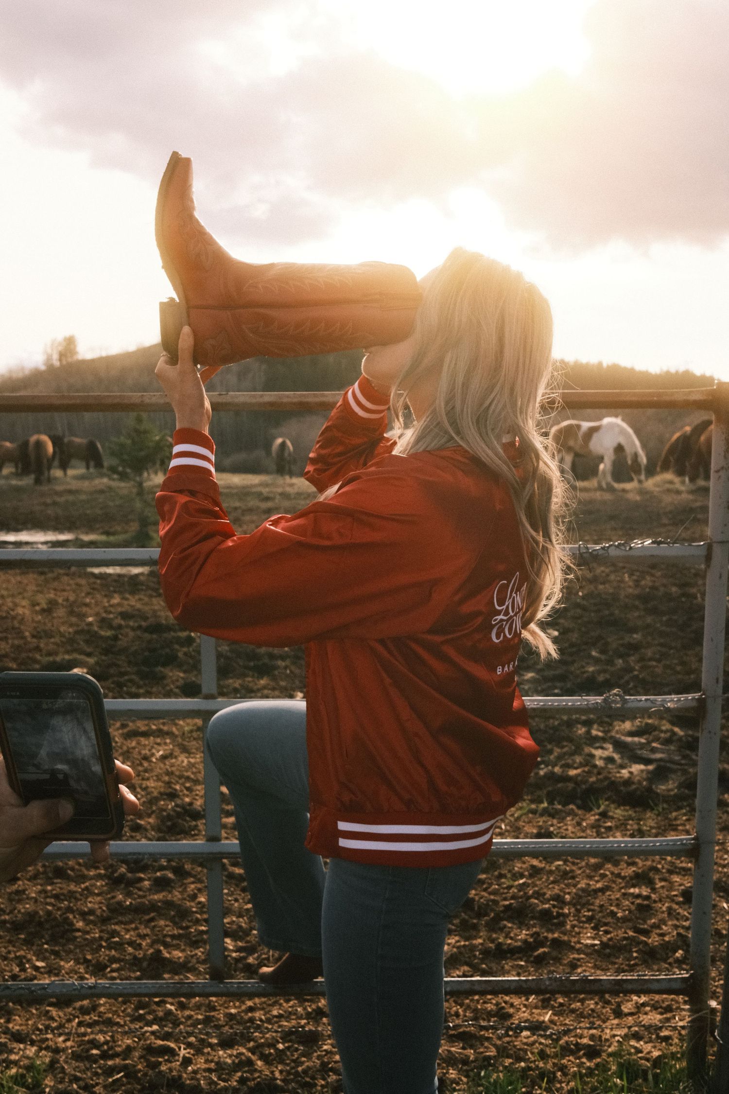 Blonde woman in red varsity jacket holding cowboy boot at sunset near horse pasture fence.