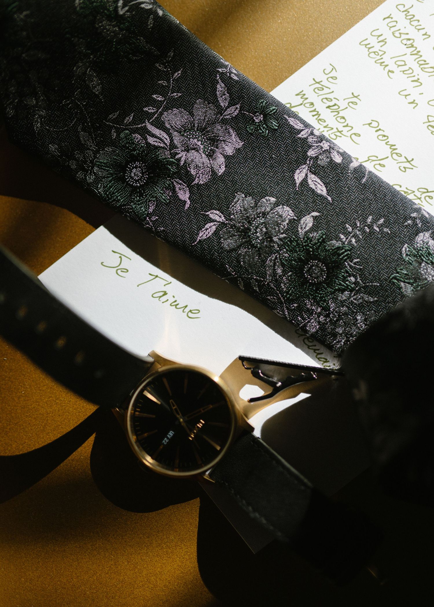 Floral patterned black tie with watch and handwritten note in moody lighting.