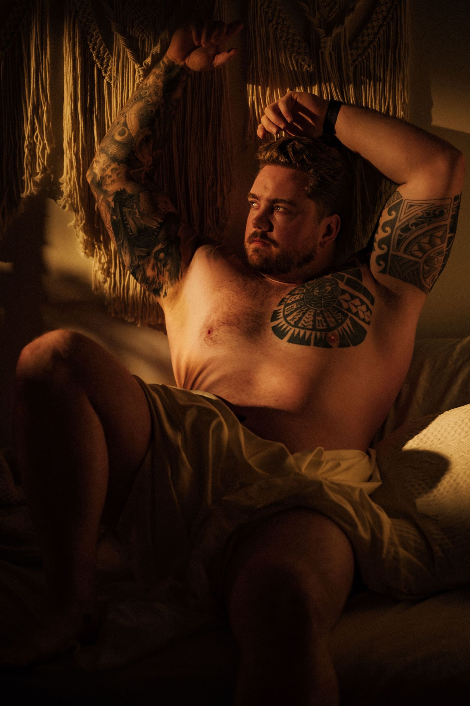 Dramatic portrait of tattooed person in moody lighting against macrame wall hanging.