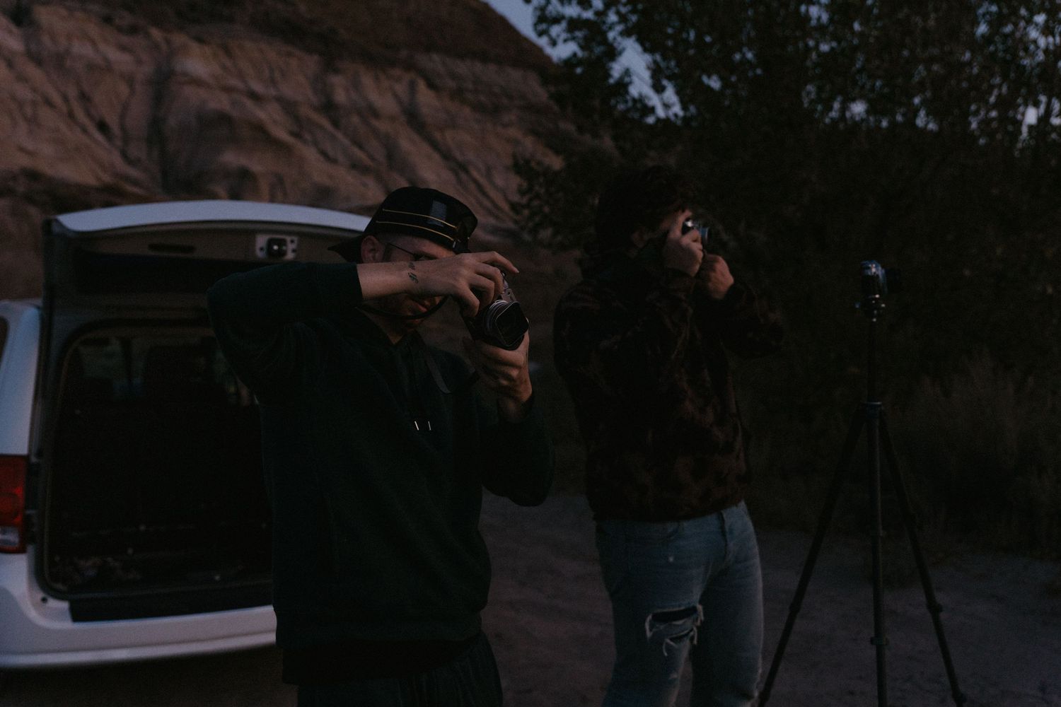 Two photographers shooting with cameras on tripods near a car at dusk with rocky cliffs in the background.