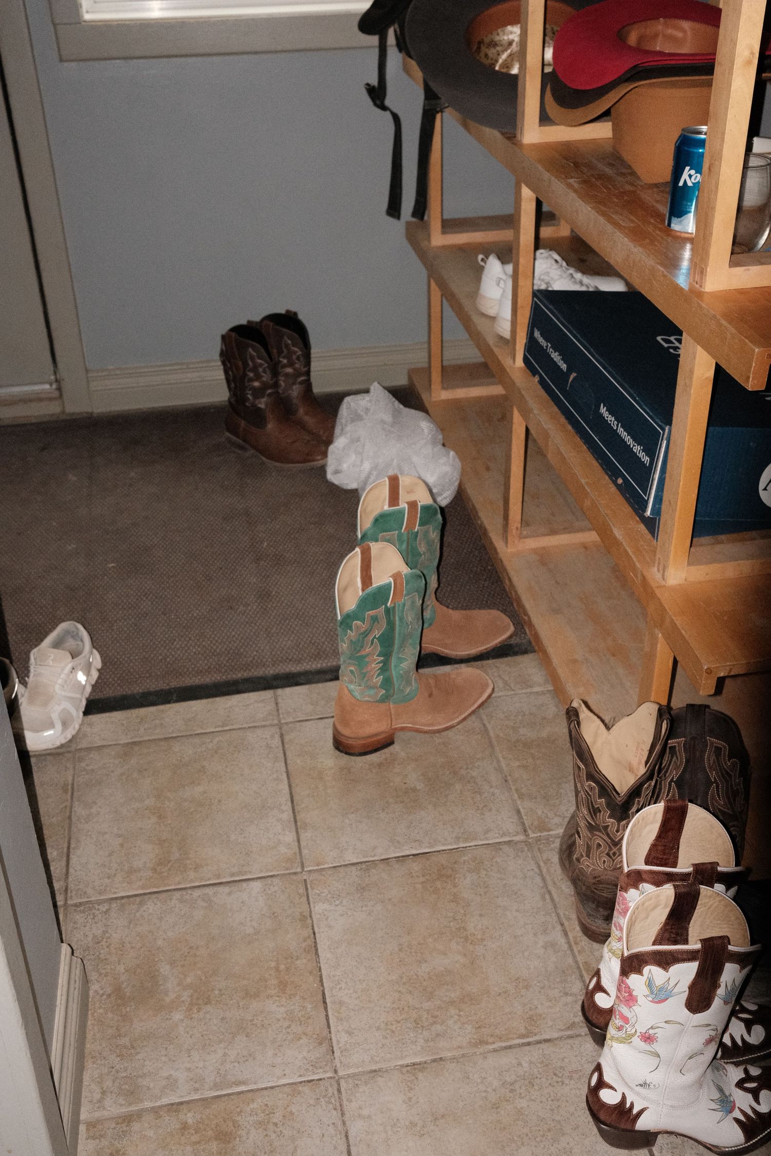 Wooden shoe rack with cowboy boots, sneakers, and boxes in a tiled entryway closet.